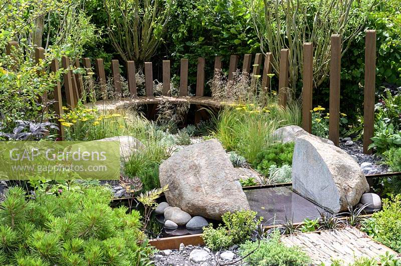 Natural rock and stone, with green foliage groundcover planting. Wooden curved bench and  corten steel reflective pool with Pinus mugo,  Deschampsia cespitosa, Achillea 'Moonshine', Ophiopogon planiscapus 'Nigrescens' -Through Your Eyes Garden - RHS Hampton Court  Palace Garden Festival 2019  - Design: Lawrence Roberts and William Roobrouck -  Sponsors: Kebony, CED Stone, R and G Metal Products, William's Art and Design, Practicality Brown.