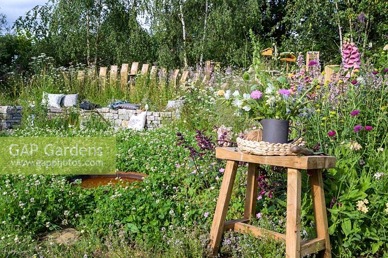 Cut flowers on old wooden stool. Cushions on curved dry stone bench backed with timber palisade surrounded by flowering perennials, water bath, clover lawn - The BBC Spring Watch Garden - RHS Hampton Court Festival - Design: Jo Thompson in consultation with Kate Bradbury