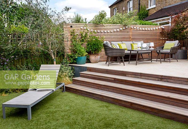London contemporary garden patio with wooden steps leading up to modern garden sofa, chairs and table on patio with cedar batten trellis fencing behind. In foreground a modern sun lounger sits on artificial lawn.