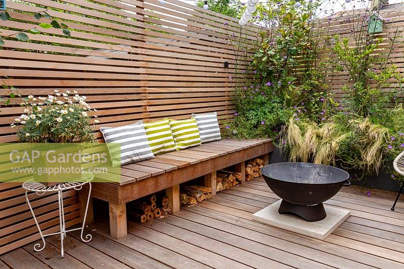 London contemporary garden -  lower wood deck area with  wooden seat and cushions. In the centre of the deck is a fire pit in front of a small raised border with Stipa tenuissima, Geranium johnsons blue, Magnolia grandiflora