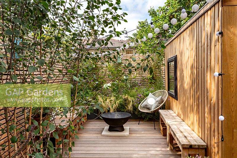 London contemporary garden -  lower wood deck area framed by a Betula utilis with  wooden seat and cushions. In the centre of the deck is a fire pit in front of a small raised border with Stipa tenuissima, Geranium johnsons blue, Magnolia grandiflora. To the right is a garden room or gym.
