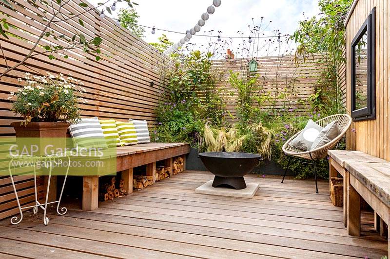 London contemporary garden -  lower wood deck area with  wooden seat and cushions. In the centre of the deck is a fire pit in front of a small raised border with Stipa tenuissima, Geranium johnsons blue, Magnolia grandiflora. To the right is a garden room or gym