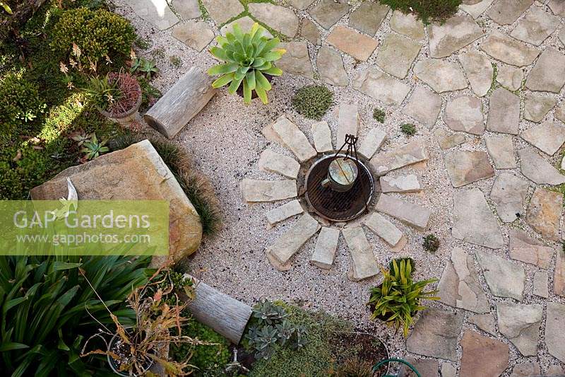 Overhead view of a firepit mulched with irrgegular flagstone paving and semi dressed rectangular sandstone blocks, with a large sandstone boulder seat, timber log seat potted plants and a garden with a variety of plants.