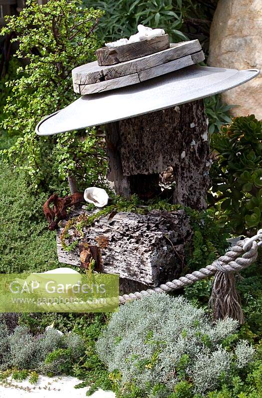 A bespoke garden feature made from wharf timbers a galvanised metal dish and ship's rope, in a garden with a dense planting of ground covers featuring Leucophyta brownii nana, Cushion Bush, with silver grey foliage. 