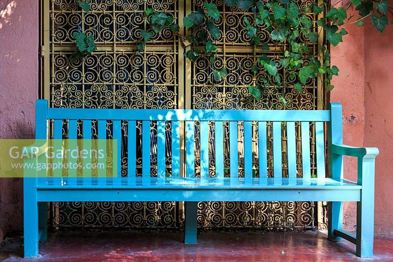 Turquoise bench in a shady corner with sunlight filtering through. Behind it a red plastered wall and ornately decorated metalwork gates.  Le Jardin Majorelle, Majorelle Garden, Marrakech