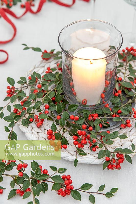 Pillar candle in storm lantern surrounded by white wicker matt and Cotoneaster berries and foliage.