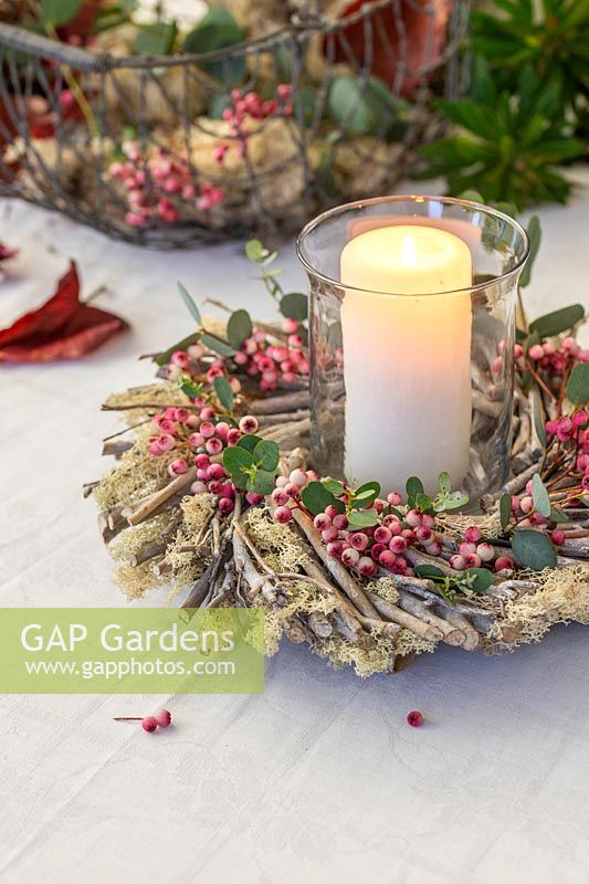 Table decoration of pillar candle in storm glass surrounded by wooden wreath decorated with pink Sorbus - Rowan - berries amd Eucalyptus foliage
