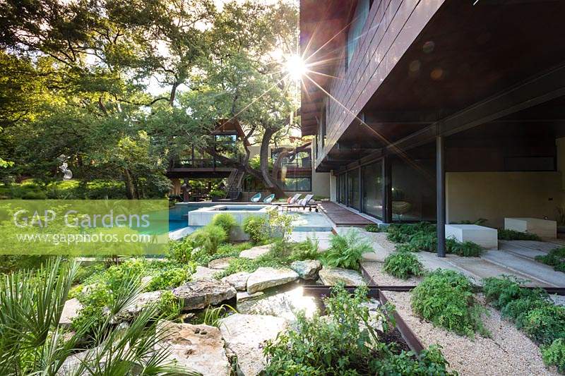 A mixture of different surfaces: rocks, gravel, pavers and decking near a contemporary house, with swimming pool and woodland garden beyond