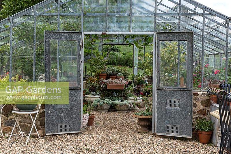 Traditional nursery glasshouse with double doors open for ventilation, showing a collection of potted plants, outdoor tables and chairs, the ground is mulched with pebbles