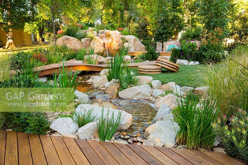 A natural looking stream and waterfall constructed from large boulders and small rocks, next to a small timber deck, interplanted with a variety of flowering and strappy leaved plants, featuring a low arched timber bridge and a small lawn area with circular seagrass cushions.