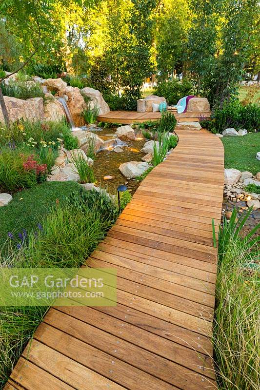 A timber boardwalk over a naturalistic stream leading to a seating area, surroundings planted out with a variety of grasses, shrubs and trees.
