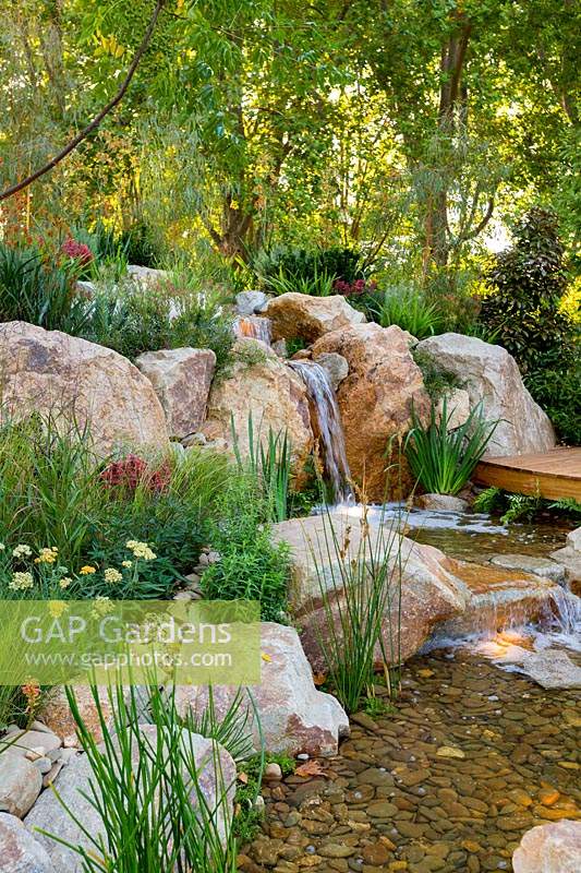 A waterfallconstructed from large boulders and small rocks, interplanted with a variety of flowering and strappy leaved plants 
