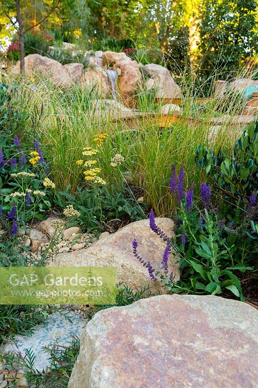 Rockery style planting, with a mixture of  grasses, ground covers and flowering herbaceous plants growing in amongst rustic sandstone rocks and river stones.