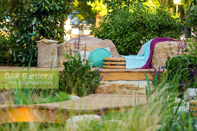 A timber deck with cushions, throws and seagrass seat, surroundings interplanted with a variety of shrubs, flowering and strappy leaved plants
