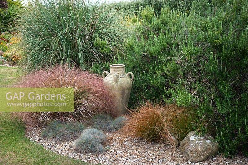 Gravel dry border with urn surrounded by plants including Anemanthele lessoniana pheasant's tail grass - syn. Stipa arundinacea, Festuca glauca blue fescue, Carex, Cortaderia and Rosmarinus officinalis rosemary