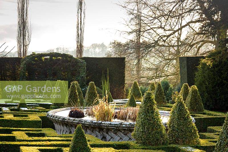 View over knot garden of clipped Buxus - Box - with variegated Box pyramids and a central raised pond, towards formal hedge with gap showing countryside beyond