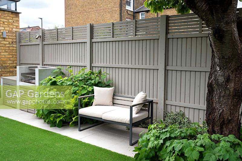 The barbeque grill and the bench with white cushions surrounded by Hydrangea and ferns by grey fence.
