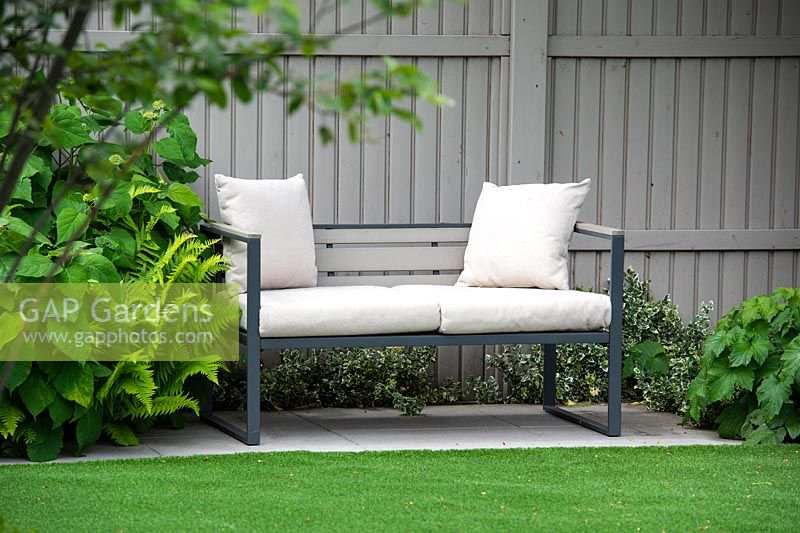 The bench with white cushions surrounded by Hydrangea and ferns by grey fence.