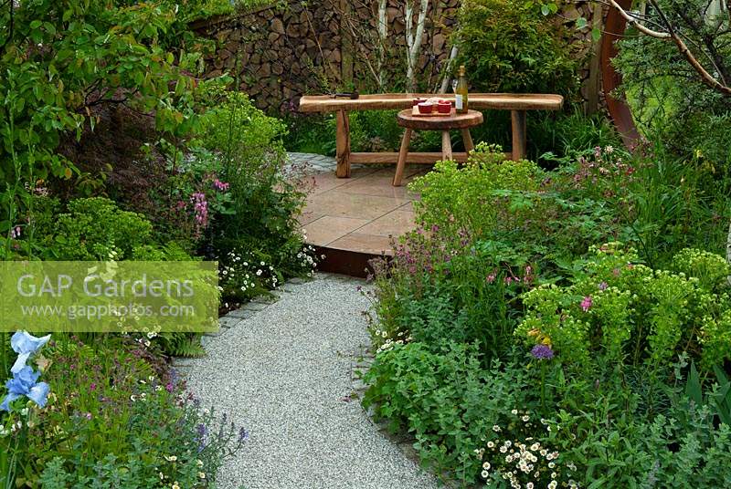 Path leading to wooden furniture on raised patio, with herbaceous borders and shrubs on either side - RHS Malvern Spring Festival 2014