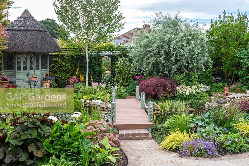 Irregular, sixth-of-an-acre town plot has a thatched  summerhouse and deck overlooking a pond edged in astilbes, arum lilies, hostas, ligularia, irises, carex and rheum. A bridge leads to mirror on back boundary. Trees: silver birch, weeping pear and Japanese maple.