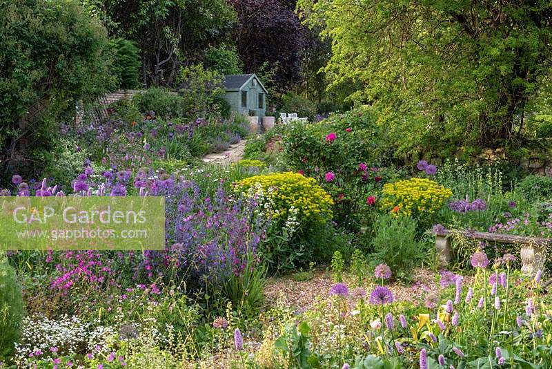 A stone bench sits in a small gravel garden edged in alliums, bistort, Euphorbia palustris, ragged robin, roses and catmint. Beyond, a paved path ascends to a summerhouse, passing a border in which flag irises and nectaroscordum star.