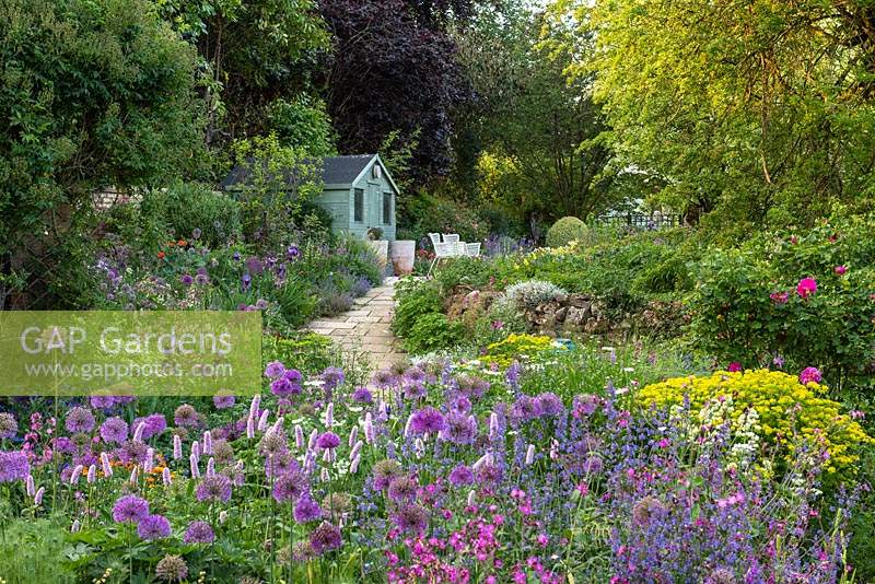 View through Allium 'Purple Sensation', Persicaria bistorta 'Superba', euphorbia, ragged robin, orlaya and catmint, to a paved path that ascends to a summerhouse, passing a border in which flag irises and nectaroscordum star.