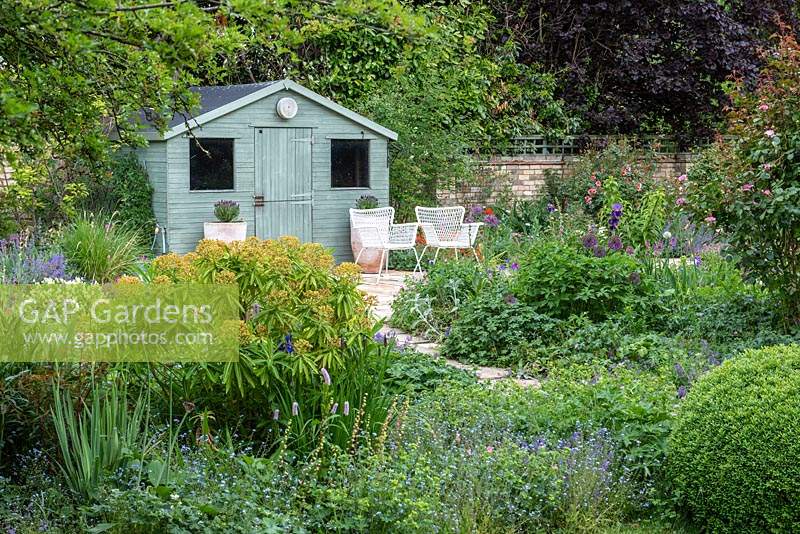 A wooden summer house and patio seating area behind deep borders planted with Euphorbia mellifera, hardy geraniums, forget-me-nots, box and Alchemilla mollis.