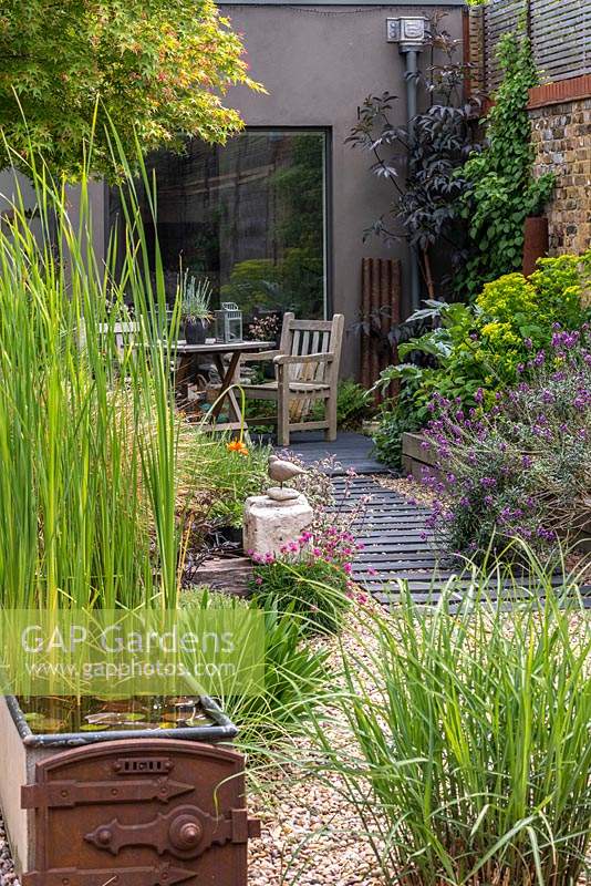 A path is made from wood off-cuts leftover from laying the wooden decks, fixed at equal intervals beneath the gravel. A quiet seating area is outside the studio, to the right a raised bed with euphorbia, black elder and mauve erysimum.