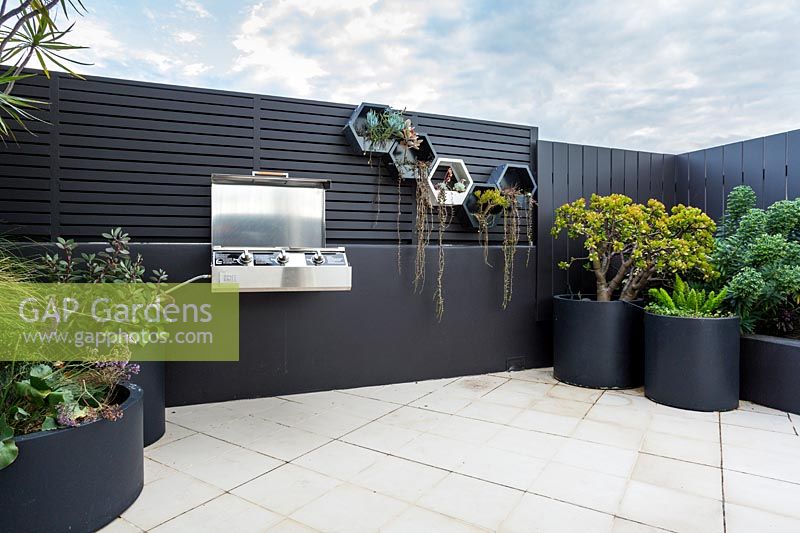 View from in doors to outside rooftop garden featuring a raised garden bed groups of grey cylindrical pots and a wall mounted barbecue.