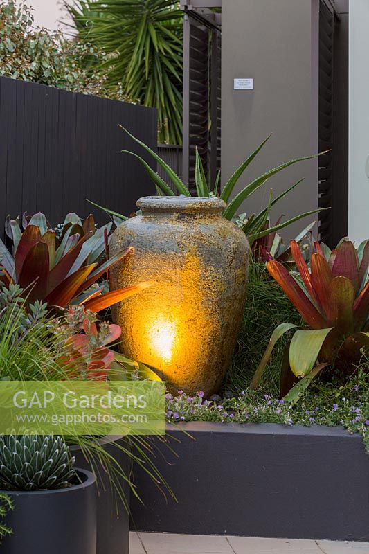 A large amphora shaped water bowl in a raised garden bed up lit by a small spotlight flanked by two large bromeliads.