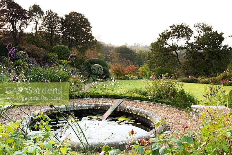 Circular pond edged with stone containing Nymphaea - Waterlily - and board for hedgehogs to climb out. Gravel path around pond offers views of garden lawn edged with borders and countryside views beyond
