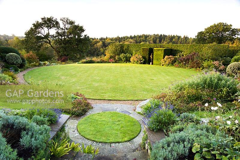 View over circular lawns edged with paving and gravel, surrounded by borders of shrubs and herbaceous perennials, with formal hedge beyond