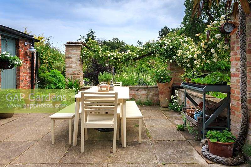 The sheltered patio featuring cream-painted table and chairs, and Rosa 'Alberic Barbier' trained along swags of thick rope