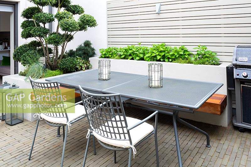 Dining area with BBQ surrounded by raised beds with Ilex Crenata 'Blondie' - Bonsai and herbs by grey wooden fence. 