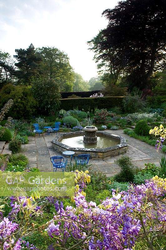 Overview of garden with central water feature fountain, blue chairs on paving between borders of shrubs and perennials. 
