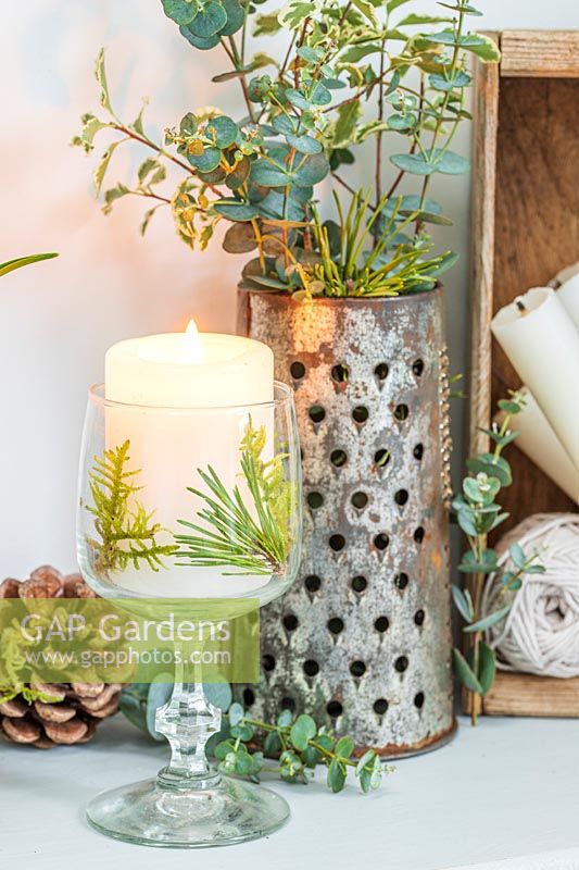 White candle in wine glass decorated with pine and moss, by grater filled with foliage