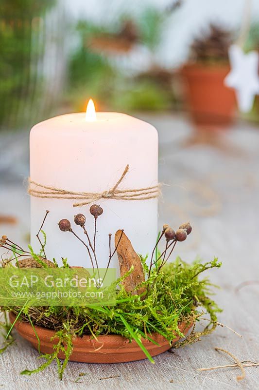 White candle on terracotta plate decorated with moss, dried lime foliage and string