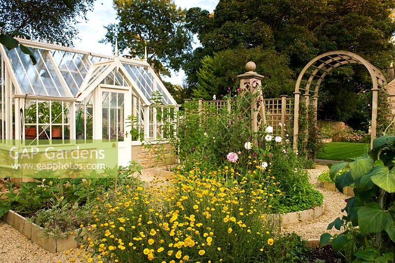Small greenhouse in vegetable garden with roses in central bed with obelisk. 