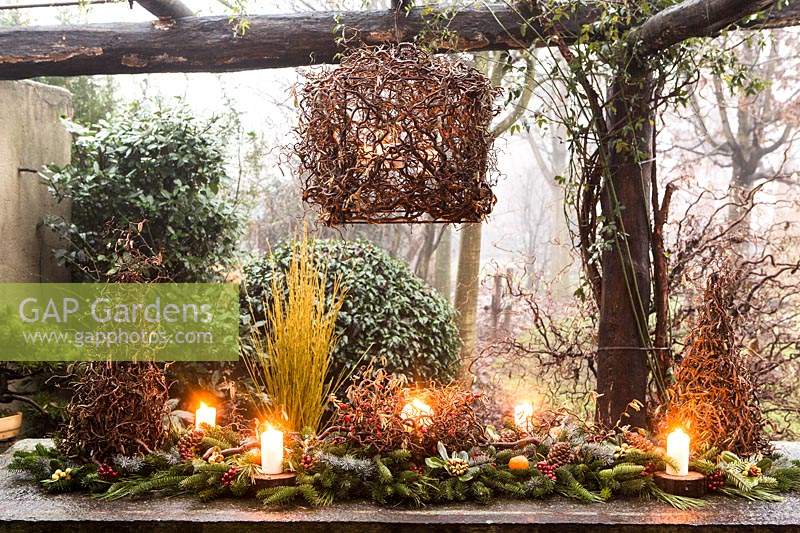 Outdoor table decoration made with Corylus avellana 'Contorta',  and branches of Abies nordmanniana, Picea pugnes 'Hopsii', Pittosporum tobira berries, pine cones, candles and mandarins