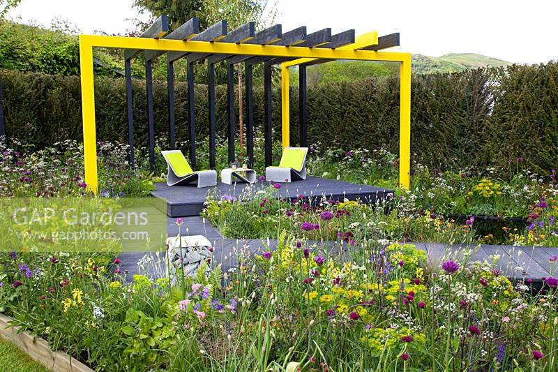 Black and yellow steel structure above black seating area and wooden platform boardwalk, surrounded by naturalistic meadow style planting in 'Urban Oasis' at RHS Malvern Spring Festival 2018