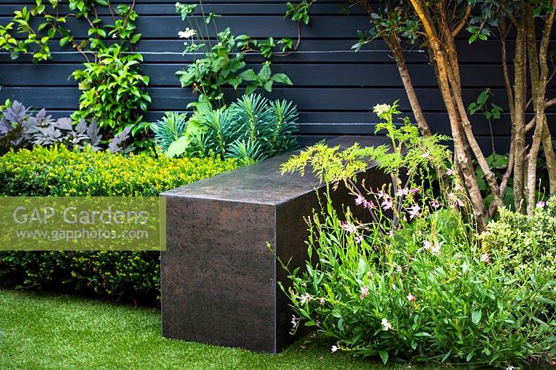 Concrete cube with porcelain cladding finished in a dark steel effect surrounded by with Gaura lindheimeriÂ 'Sparkle White', Osmanthus x burkwoodii, Taxus baccata and metal cube by black wooden fence. 
