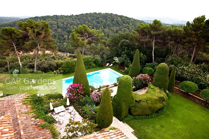 Over view of garden with seating area, large topiary forms, swimming pool and views of trees and wooded hills beyond 
