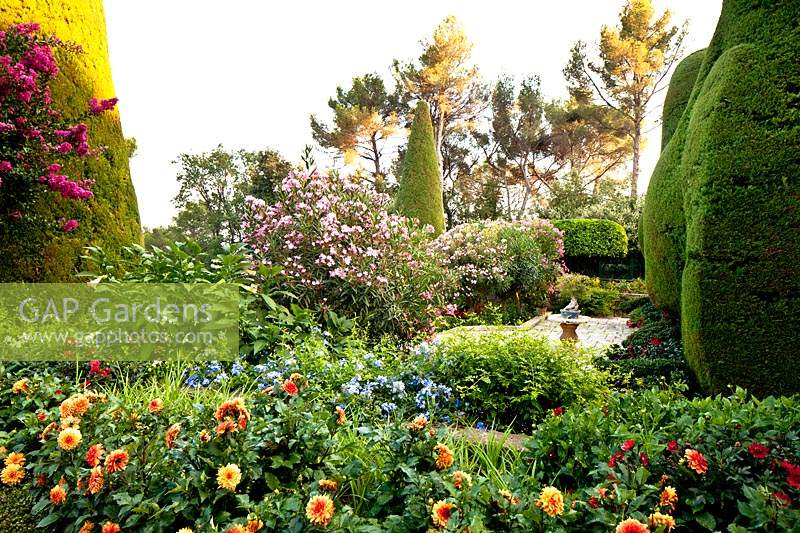 View over flower beds to paved terrace with shrubs, large topiary forms and trees beyond