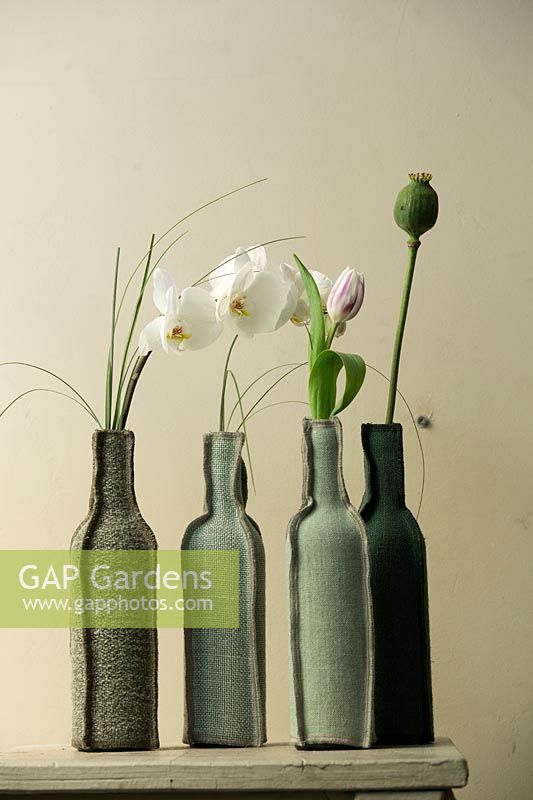 Fabric bottles containing Orchid, Tulip and poppy seed head on shelf. 