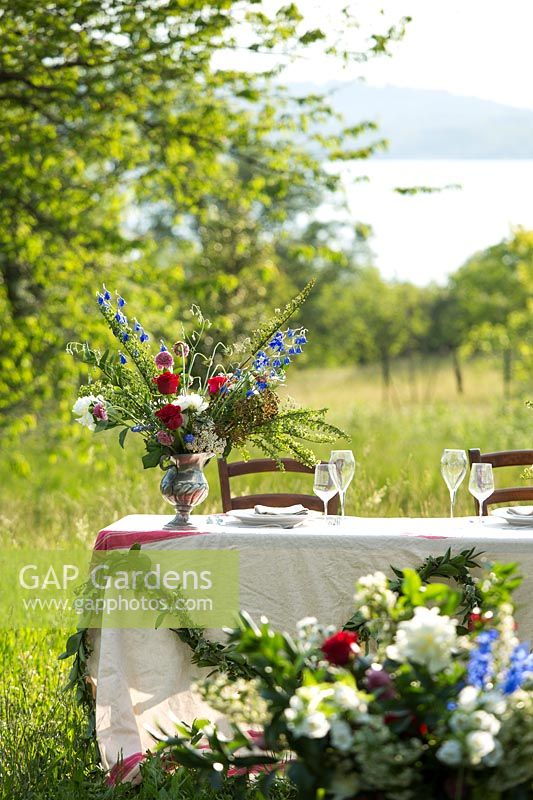 Table in countryside, with floral arrangement and foliage swag. Flowers include Delphinium, Rosa - Roses and Paeonia - Peony 