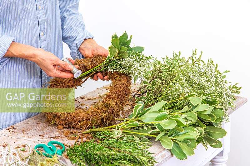 Woman using floristry wire to fix bundle of cut flowers and foliage to moss wreath form. 