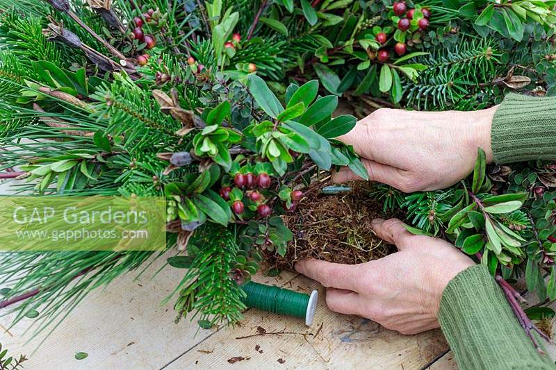 Woman using floristry wire to attach bundle of assorted foliage, stems and berries to moss wreath form