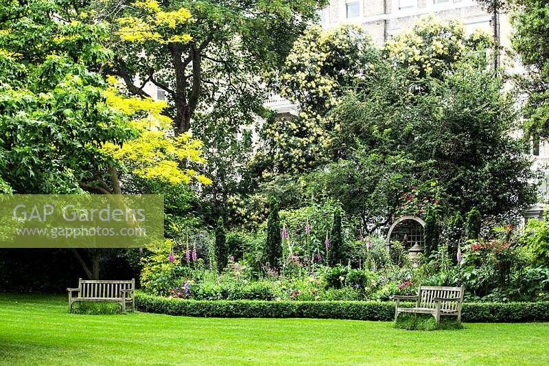 Square garden with lawn, benches, beds with trees in front of buildings beyond