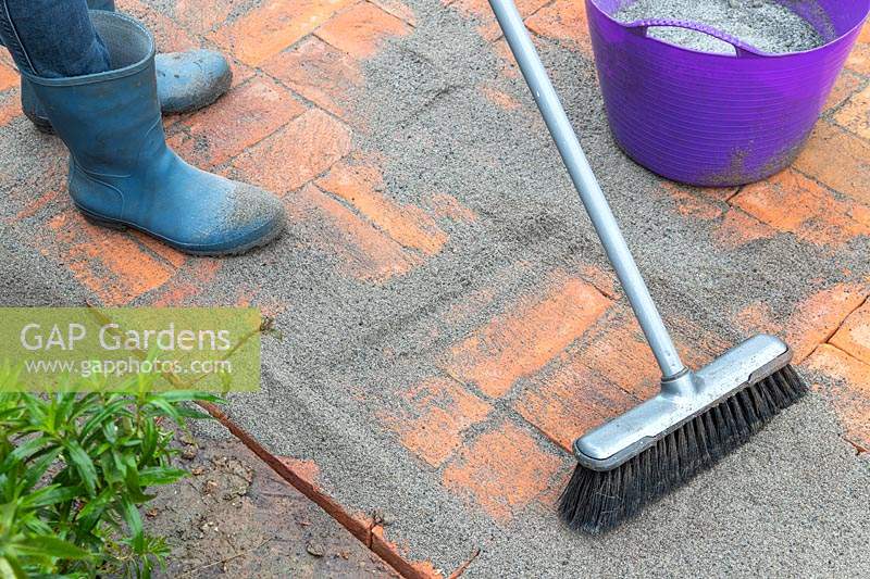 Woman using soft brush to push polymer sand into joins of newly laid brick path