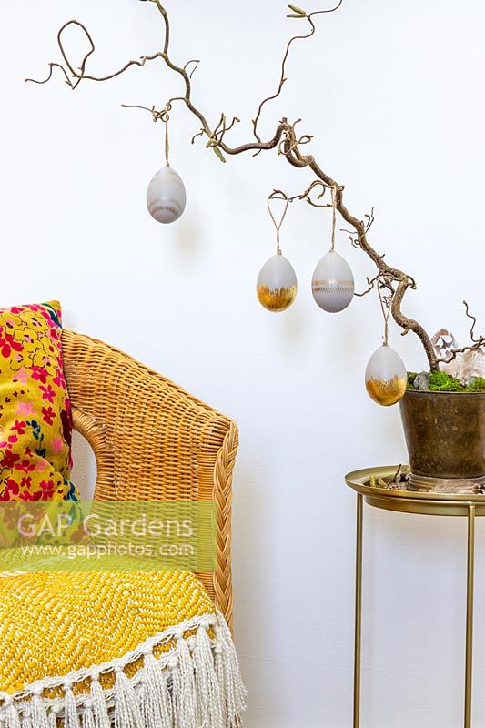 Grey and gold painted Easter Eggs hanging from twisted Hazel branch with gold pot and stand next to wicker chair